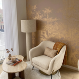 A styled interior corner featuring a cream bouclé armchair with a plaid throw pillow and blanket, next to a round wooden coffee table and a tall floor lamp, set against a beige wall with gold botanical patterned wallpaper.