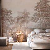 A styled bedroom featuring a large wall mural with a soft, vintage-style forest landscape in muted beige and brown tones. The room includes a low upholstered bed with neutral bedding, a rustic wood side table with glass lamps, a white vase with dried stems, and a small gray side stool on a light rug.