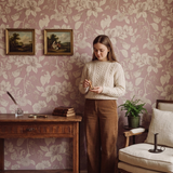 A woman stands in a vintage-style room with pink floral wallpaper, wearing a cream cable-knit sweater and brown wide-leg trousers. She is positioned next to a wooden desk, framed paintings, a potted plant, and a cream-colored armchair.