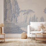 A styled living room featuring a large wall mural with a classical landscape scene of cypress trees and a river, styled with a white sofa, wooden coffee tables, and a beige rug.
